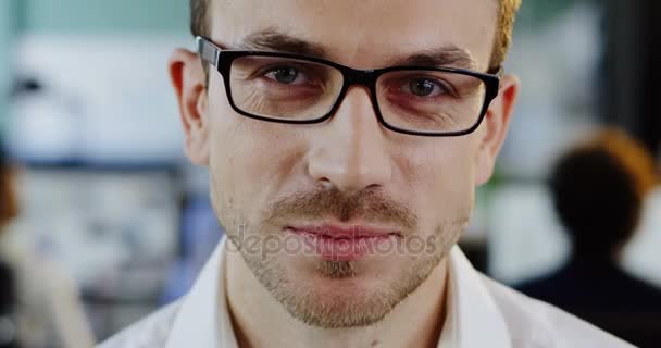 Close up de homem bonito caucasiano em óculos olhando e sorrindo sinceramente direto para a câmera. O fundo turvo da sala de escritório. Retrato tirado — Vídeo de Stock