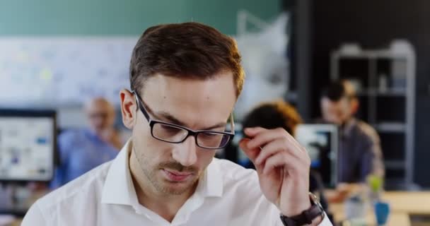 Primer plano de un joven guapo caucásico con gafas mirando a la cámara, arreglando sus gafas y sonriendo. El fondo borroso de la oficina. Un retrato. En interiores — Vídeo de stock