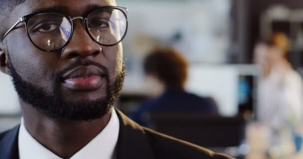 Gros plan de jeune homme attrayant afro-américain dans des lunettes regardant sur le côté et souriant directement dans la caméra. Le bureau flou avec le fond des travailleurs. Portrait. Intérieur — Video