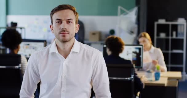 Retrato de homem jovem atraente na camisa branca que olha para a câmera e o escritório borrado com funcionários em computadores atrás. Interior — Vídeo de Stock