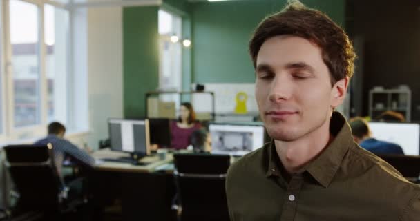 Portrét mladé atraktivní muž otočil hlavu k fotoaparátu v moderní kanceláři. Pracujících na pozadí. Detailní záběr. — Stock video