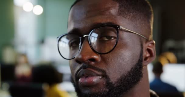 Närbild av unga attraktiva afroamerikanska manliga kontorist i glasögon att vrida huvudet till kameran och leende på urban kontoret. Arbetande människor på den suddig bakgrunden. Porträtt — Stockvideo
