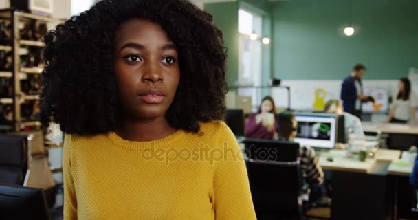 Primer plano de la joven afroamericana rizada girando la cabeza a la cámara y sonriendo sinceramente en la oficina urbana. Gente trabajadora en el fondo. Retrato — Vídeos de Stock