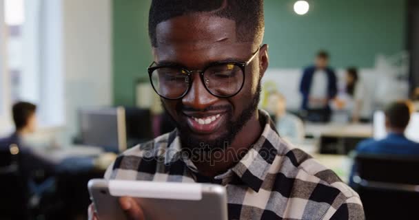 Primer plano del joven guapo afroamericano con gafas trabajando en la tableta y sonriendo a la cámara en la oficina moderna. Gente trabajadora en el fondo borroso. Retrato plano — Vídeo de stock