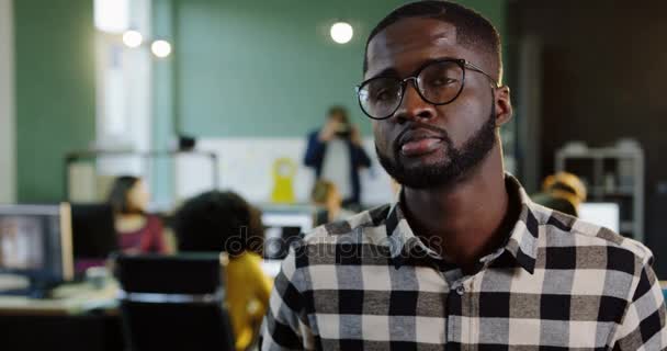 Retrato de un joven afroamericano con gafas girando la cabeza hacia la cámara y sonriendo en la oficina moderna. Gente trabajadora en el fondo borroso. De cerca. — Vídeos de Stock