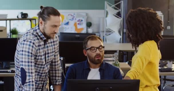 Kaukasischer Mann mit Brille, der einer jungen Frau mit schwarzem Lockenkopf und einem anderen kaukasischen jungen Mann etwas vorschlägt. Business-Projektteam. in der Nähe des Computers in den schönen Büroräumen. — Stockvideo