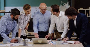 Belgeler, çizelgeler, grafikler ve finansal raporlar ile bir masa etrafında duran ve neşeyle onların patron hakkında başlangıç ile danışmanlık iş işçilerin genç grup. Modern ofis alanı içinde