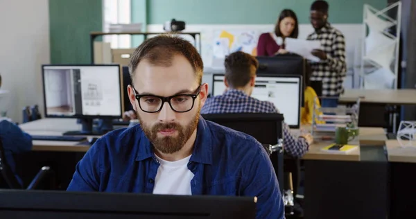 Close up of young handsome Caucasian man in glasses working on the computer and thinking in the modern comfortable office space. Office workers on the blurred background. Portrait