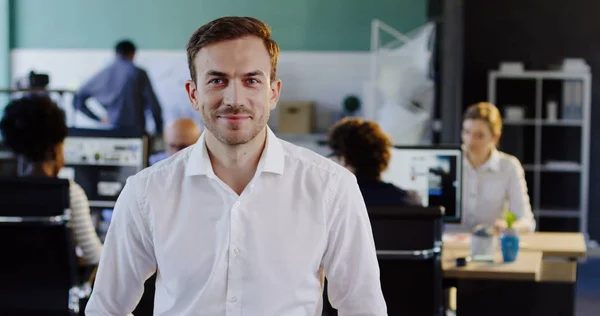 Portrait of attractive young man in white shirt looking into the camera and the blurred office with workers at computers behind. Indoor