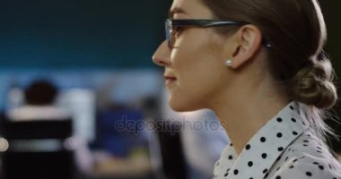 Ofis alanı arkasında, daha başından kameraya dönüm ve içtenlikle gülümseyen bakarak gözlük güzel kadının kapatın. Akşam geç. Kapalı. Portre