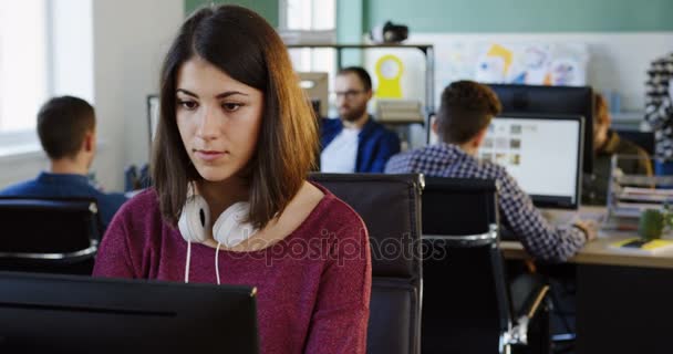 Retrato tiro de mulher atraente jovem com fones de ouvido trabalhando no computador e sorrindo no espaço de escritório moderno. Trabalhadores de escritório em segundo plano. Fechar . — Vídeo de Stock