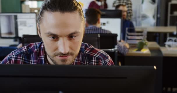 Close up de jovem caucasiano bonito em camisa xadrez trabalhando no computador e sorrindo na sala de escritório moderna. Trabalhadores de escritório no fundo turvo. Retrato — Vídeo de Stock