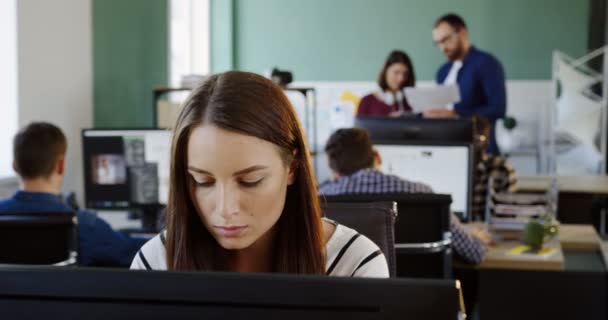 Primer plano de la joven hermosa mujer que trabaja pensativamente en la computadora en el espacio de oficina moderno. Trabajadores de oficina en segundo plano. Retrato plano — Vídeo de stock