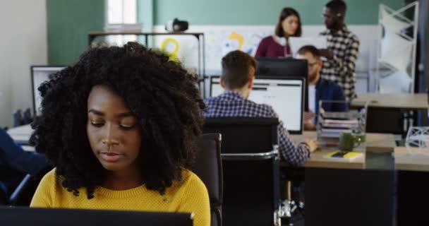 Retrato de jovem mulher afro-americana encaracolado atraente em suéter amarelo trabalhando no computador e sorrindo na sala de escritório moderna. Trabalhadores de escritório em segundo plano. Fechar . — Vídeo de Stock