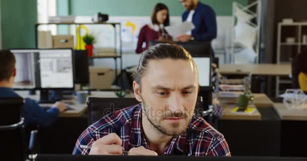 Portrait de jeune homme blanc attrayant employé de bureau en chemise à carreaux travaillant soigneusement sur l'ordinateur dans l'espace de bureau moderne. Des employés de bureau sur fond flou. Gros plan 