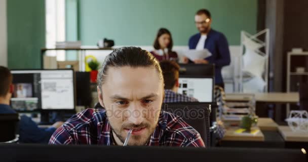 Primer plano del joven guapo trabajador de oficina caucásico con camisa a cuadros que trabaja duro en la computadora en la oficina urbana. Trabajadores de oficina en el fondo borroso. Retrato — Vídeo de stock