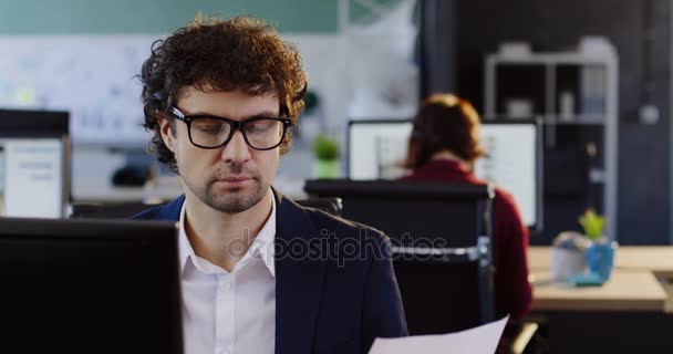 Trabajador de oficina sentado en la computadora y trabajando con documentos impresos en la moderna sala de oficina. De interior — Vídeos de Stock