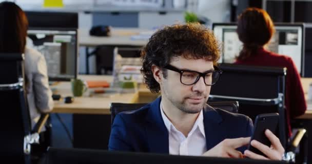 Retrato de homem atraente encaracolado em óculos rolando no telefone celular enquanto sentado em seu local de trabalho no escritório. O fundo de empregados de escritório de trabalho. Interior — Vídeo de Stock