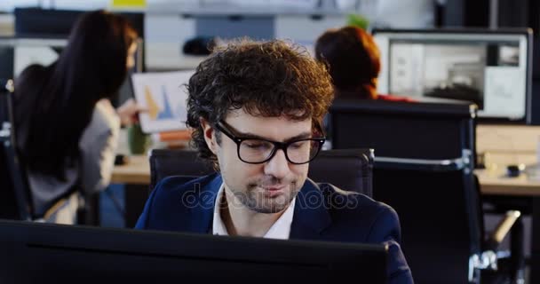 Retrato tiro de adorável empregado do sexo masculino em óculos chamando o telefone celular enquanto sentado em seu local de trabalho no escritório da grande empresa. O fundo dos trabalhadores de escritório nas suas mesas. Para dentro — Vídeo de Stock