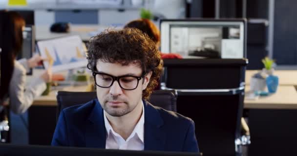 Portrét úřednice vážné atraktivní muž v brýlích, pracovat na počítači a mluví o mobilní telefon v moderní kanceláři. Rozmazané prostor s pracovníky na počítači v pozadí — Stock video