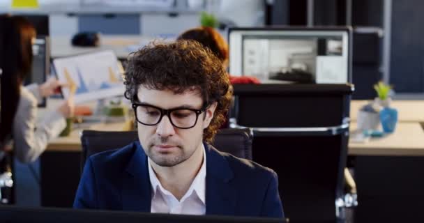 Retrato de un hombre serio y atractivo oficinista en gafas trabajando en el ordenador y hablando en el teléfono móvil en la oficina moderna. El espacio borroso con los trabajadores en las computadoras en el fondo — Vídeo de stock