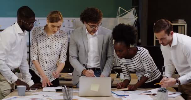 Mladý podnikatelský tým multietnických kancelářských pracovníků, který kontroluje data a pracuje na novém startupním projektu, zatímco stojí kolem stolu s dokumenty, grafy, grafy a finančními zprávami — Stock video