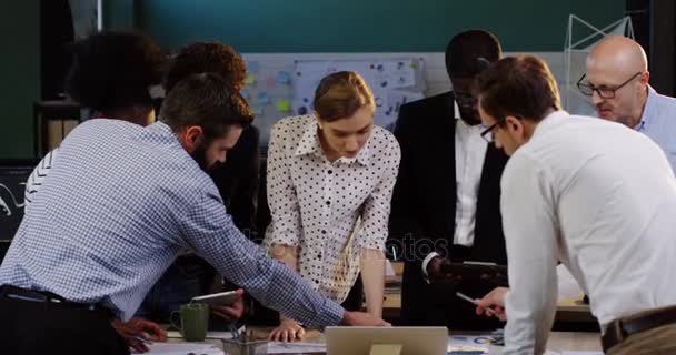 Equipo de carreras mixtas de compañeros de trabajo apoyados en una mesa de conferencias mientras consultan sobre su proyecto empresarial y sus planes financieros. Dentro de la oficina urbana — Vídeos de Stock