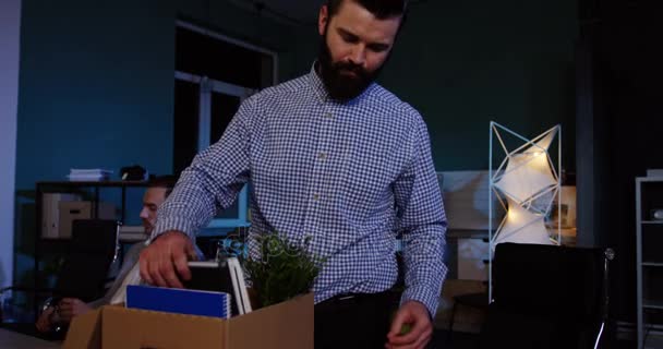 Um jovem caucasiano com barba a arrumar as coisas dentro da caixa do escritório à noite, porque perdeu o emprego. Interior — Vídeo de Stock