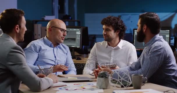 Four businessmen sitting at the table and discussing their future plans, than shaking hands in the comfortable office late in evening. Indoors. Table with documents and charts — Stock Video