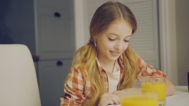 Carina ragazza bionda seduta al tavolo della cucina con un bicchiere di succo di frutta e sua madre le porta un piatto di muffin. Figlia che abbraccia sua madre. Dentro. — Video Stock