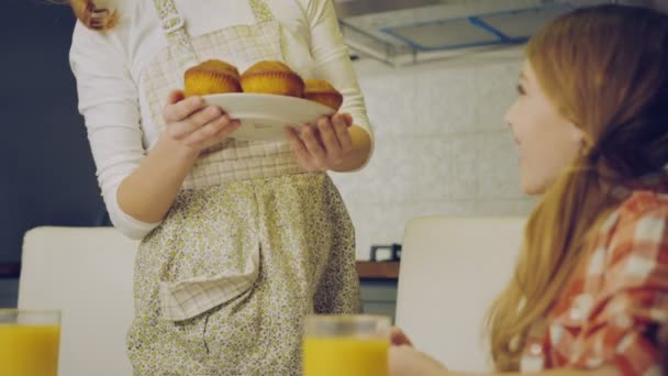 Όμορφη μητέρα στην ποδιά φέρνοντας και θέτοντας σε muffins πίνακα κουζινών για την πλάκα και την πανέμορφη κόρη που αγκάλιαζε. Μαμά, φιλί ένα κορίτσι στο μέτωπο. Σε εσωτερικούς χώρους — Αρχείο Βίντεο