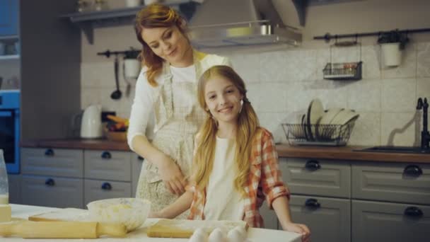 Retrato tiro da bela mãe abraçando sua filhinha bonita e ambos sorrindo a câmera na cozinha na mesa enquanto assando. Para dentro — Vídeo de Stock