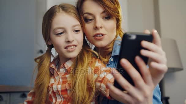 Primer plano de la hermosa hija y madre sentadas una junto a la otra y haciendo selfies en el teléfono inteligente negro en la habitación en casa. En interiores. Retrato — Vídeos de Stock