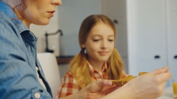 Närbild på vackra mor sprida jordnötssmör på brödet med en sked för hennes dotter medan hon tittar på henne i köket. Inuti — Stockvideo