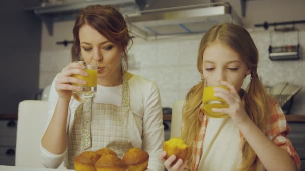 Retrato da mãe atraente e sua linda filha adolescente sentada na cozinha, comendo muffins, bebendo suco e abraçando. Interior — Vídeo de Stock