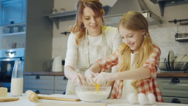 Симпатичная девушка-подросток разбивает яйцо о стеклянную миску с доу внутри и ее мать наблюдает, как она делает это и контролирует приготовление пищи. Портретный снимок . — стоковое видео