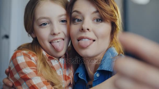 Retrato de la hermosa mujer con hija pequeña haciendo selfies en el teléfono inteligente, mostrando sus lenguas, abrazando y riendo. De cerca. Adentro. — Vídeo de stock