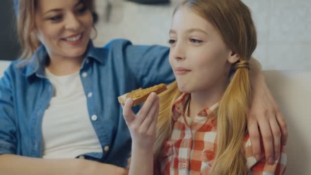 Close up da menina bonita adolescente mordendo e mastigando um pão com manteiga de amendoim perto de sua mãe sorridente. Tiro de retrato. Para dentro — Vídeo de Stock