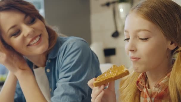 Primer plano de la linda chica adolescente disfrutando de un sándwich con mantequilla de maní y su madre mirándola, que abrazándola y besándola. Un retrato. En interiores — Vídeo de stock