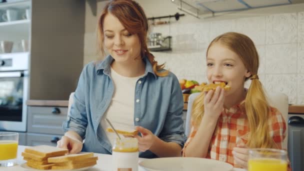 Retrato de la hermosa madre haciendo sándwiches con mantequilla de maní y su encantadora hija comiéndolos con una sonrisa. De interior — Vídeos de Stock