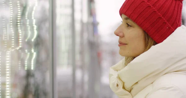 Close up of the beautiful blonde woman in warm winter coat and red hat coming closer to the window case and looking into it. Outdoor. Portrait shot