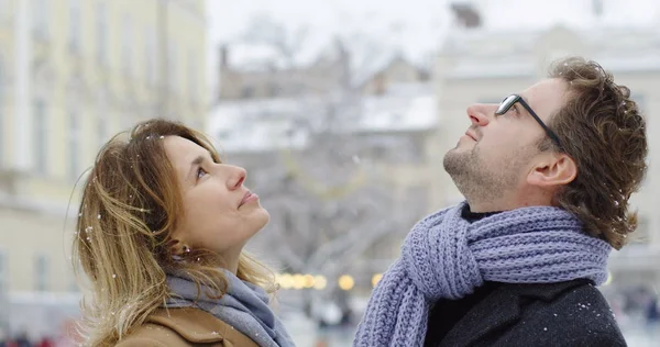 Primer plano de la feliz pareja de mediana edad mirando hacia arriba y riendo mientras está nevando en el medio de la bonita ciudad en el día de invierno. Al aire libre. Retrato plano — Foto de Stock