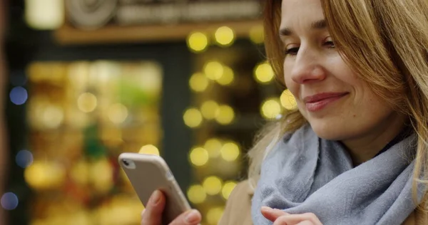 Portrait of the charming womans face of middle age looking happy of what she seeing on the smartphone screen. The Christmas decorated shops with lights on the background. Outdoor. Close up