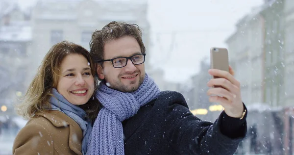 Retrato de la atractiva pareja de mediana edad haciendo selfies en el teléfono inteligente en medio de la bonita plaza de la ciudad mientras nieva en el día de invierno. Exterior . — Foto de Stock