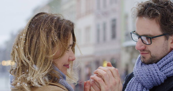 Close up of the lovely middle aged couple standing close, looking at each other gently and man warming womans hands on the snowy day in town. Portrait shot. Outdoors.