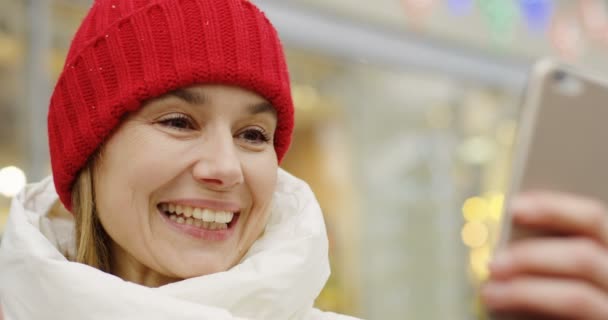 Retrato de la mujer rubia de mediana edad en el sombrero rojo videochat en el escaparate borroso con las luces de Navidad de fondo. Al aire libre. De cerca. — Vídeo de stock
