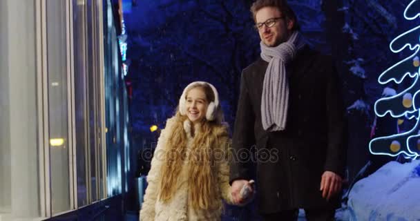 Όμορφη νεαρή κοπέλα περπάτημα χέρι-χέρι με τον πατέρα της κατά μήκος τις βιτρίνες των καταστημάτων για τα Χριστούγεννα διακόσμηση δρόμο το βράδυ. Εξωτερικό — Αρχείο Βίντεο