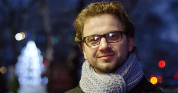 Portrait shot of the handsome man in glasses and grey scarf turning his head to the camera and smiling in it. Christmas night in the street. Outside. Close up — Stock Video