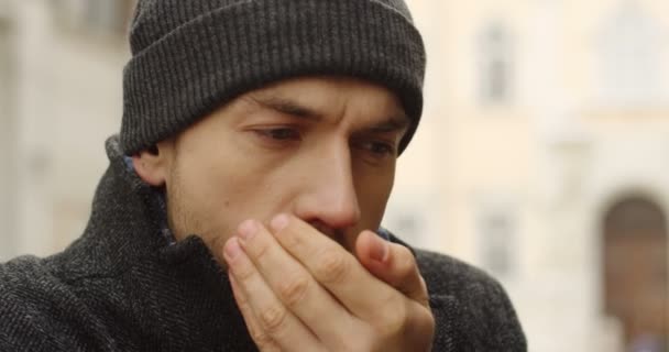 Retrato del hombre caucásico con un sombrero negro calentando sus manos en el frío invierno. Fondo borroso. De cerca. Al aire libre — Vídeo de stock