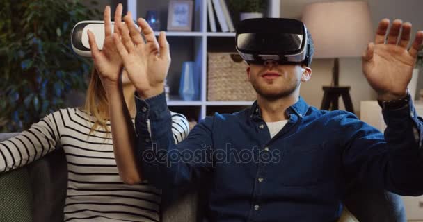 Coppia giovane seduta sul divano in salotto con occhiali VR e auricolare VR, scorrimento e nastro adesivo in aria. Ora di sera. Chiudete. Ritratto. Dentro. — Video Stock